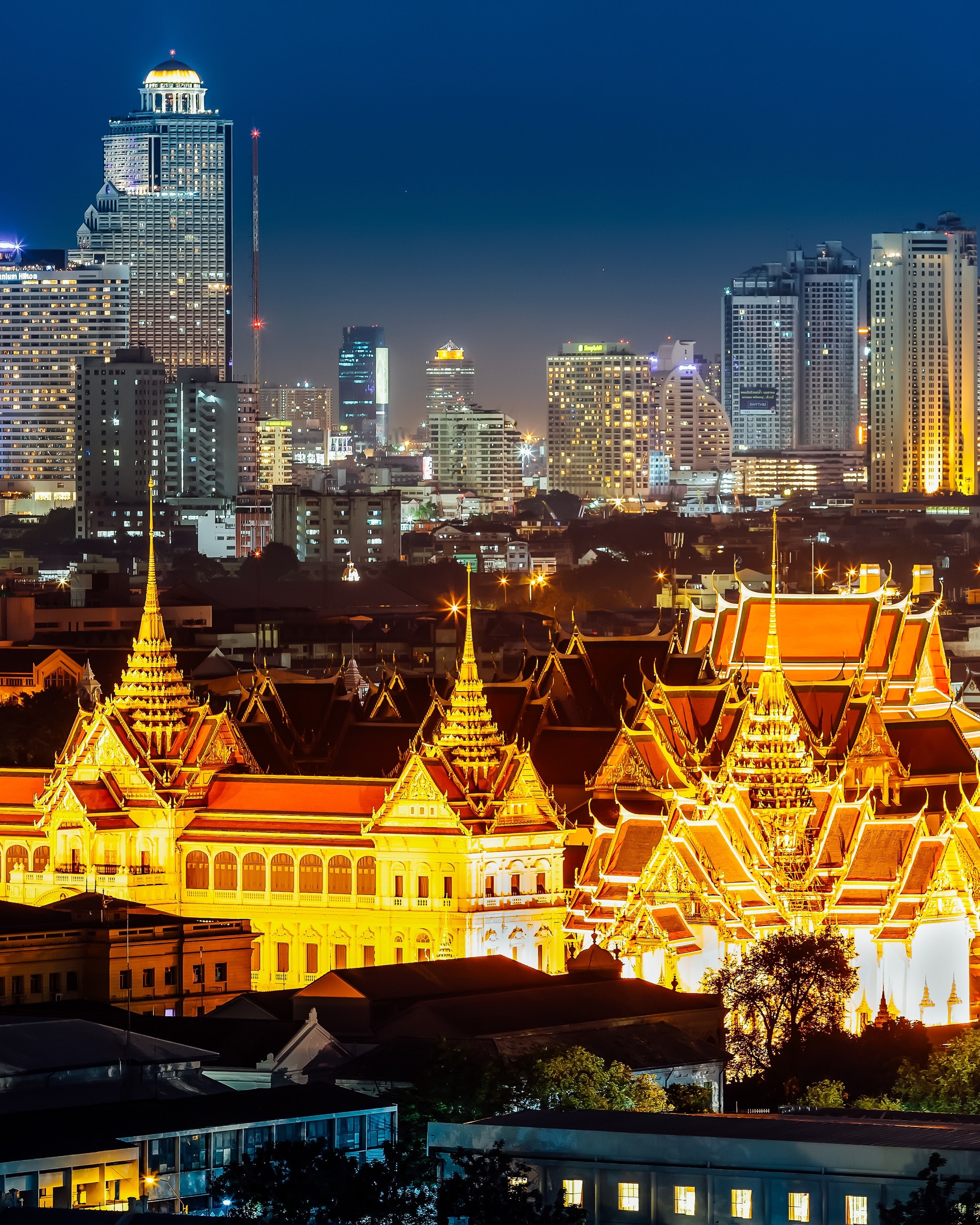The Grand Palace lit at night, with the Bangkok skyline behind it